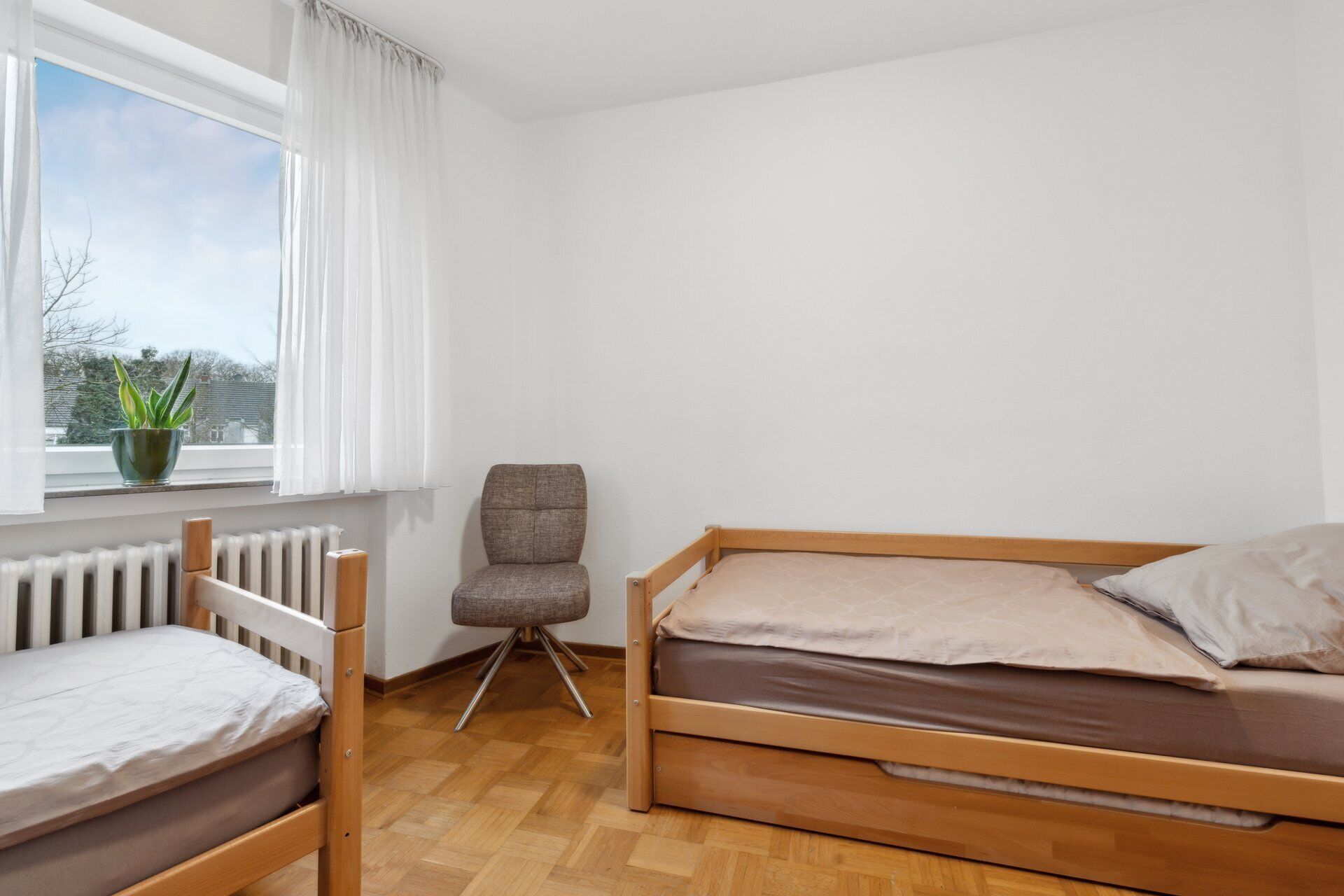 A small, well-lit room with a wooden bed, a chair, a radiator, a window with curtains, and a potted plant.