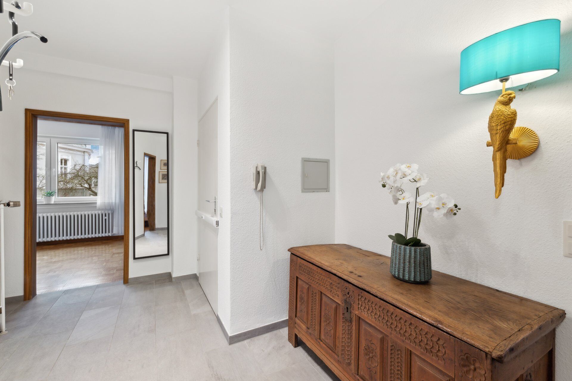 A hallway with a wooden cabinet, a wall-mounted lamp, and a mirror reflecting a room with a window.
