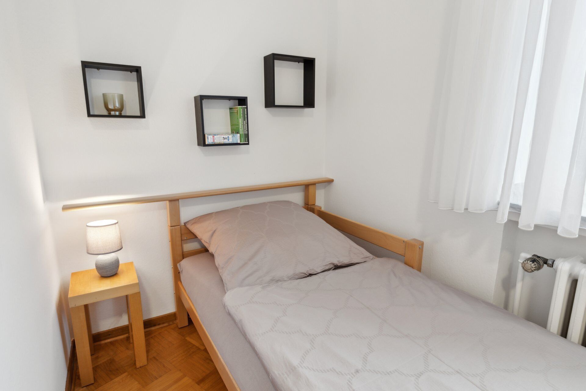 Een kleine, verzorgde slaapkamer met een enkel bed, een nachtkastje met een lamp, en drie ingelijste decoraties aan de muur.