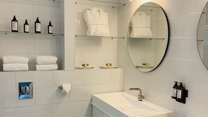 Une salle de bain moderne avec des carreaux blancs, des toilettes, deux lavabos, un miroir et des étagères contenant des articles de toilette.