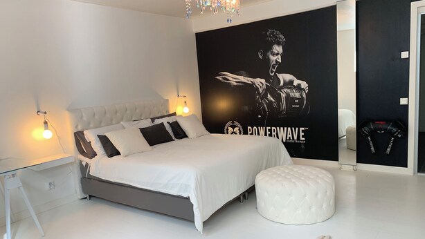 Une chambre moderne avec un grand lit, un pouf blanc, un tapis en fourrure, un miroir fixé au mur et une affiche représentant un boxeur.