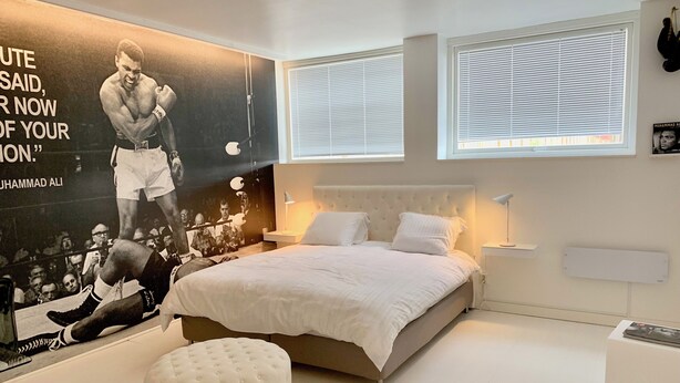 Une chambre avec un grand lit, un pouf rond, une affiche au mur et une fenêtre avec des stores.