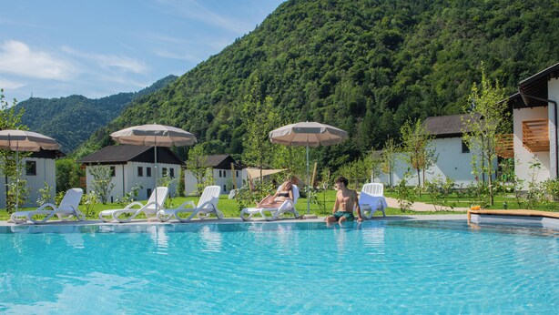 A swimming pool with lounge chairs and umbrellas, surrounded by buildings and a forested hillside. - In the heart of the Ledro Valley, where nature sets the pace. (Trentino-Alto Adige)