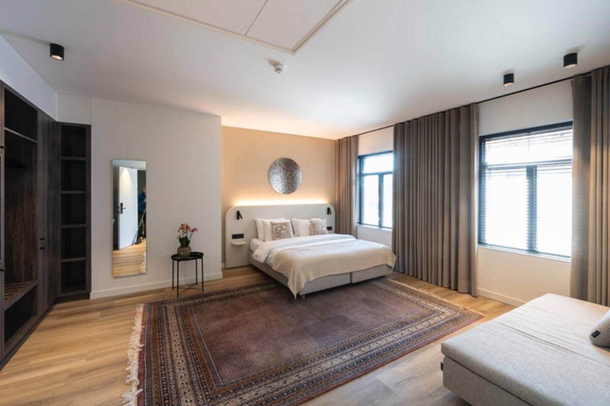 A modern bedroom with a large bed, a small side table with a vase, a mirror, a large window with curtains, and a built-in wardrobe.