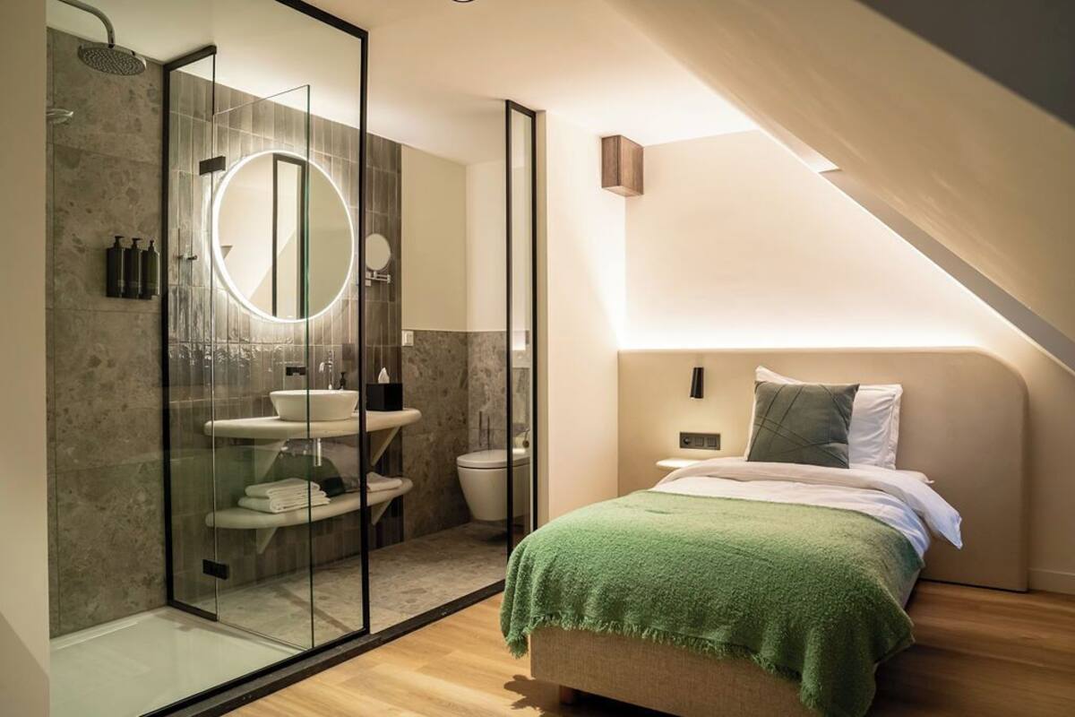A modern bedroom with a glass-enclosed shower, a round mirror, a bed with a green blanket, and a small bedside table.
