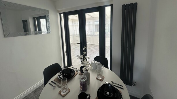 A dining area with a round table, chairs, a chandelier, and a view through large windows. - Housesleeps6-freeparking-wifi-contractors-garden (Billingham)