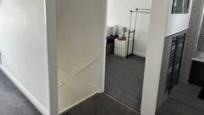 A hallway with a white door, a small bedside table with a lamp, and a mirror on the wall. - Housesleeps6-freeparking-wifi-contractors-garden (Billingham)