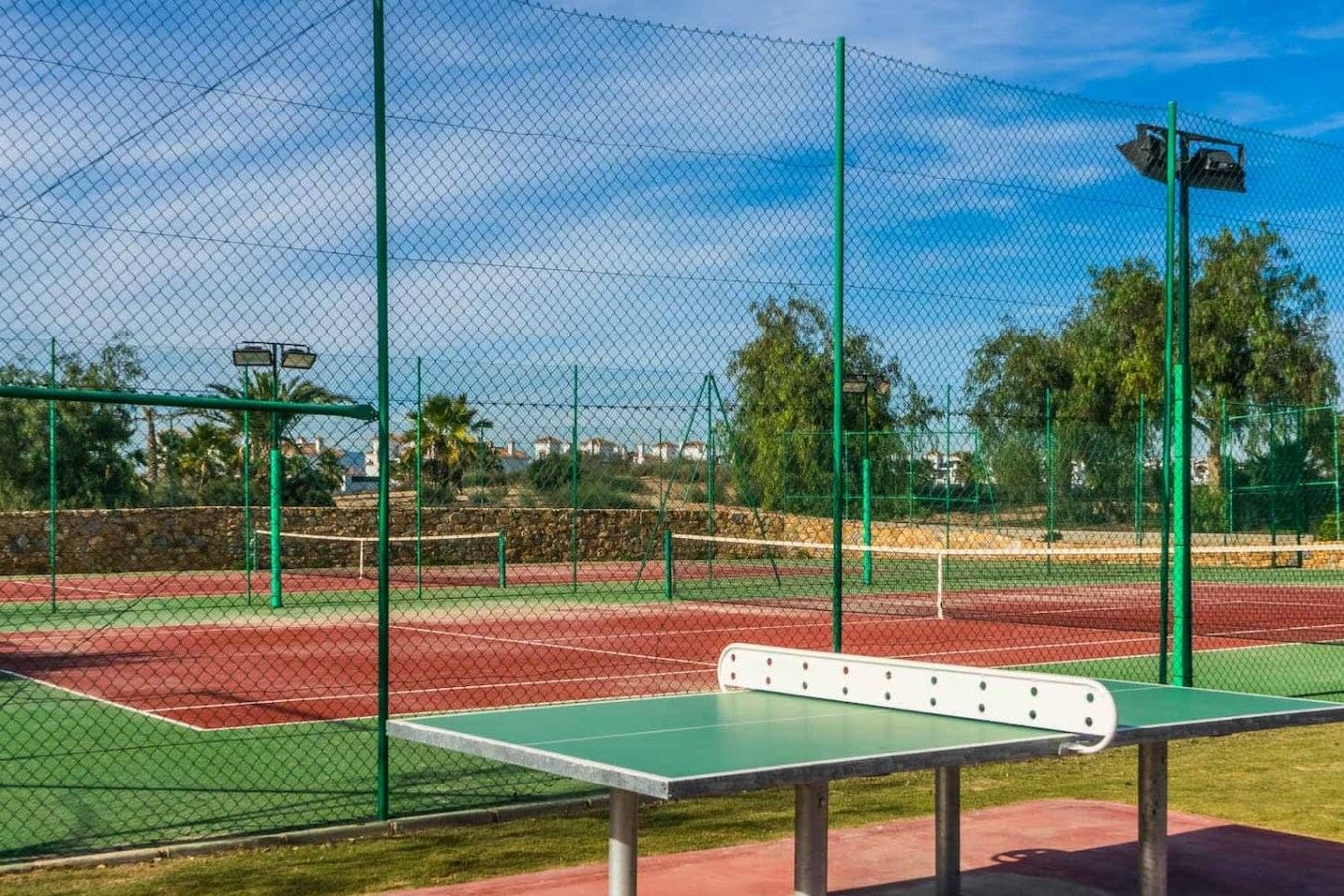 An outdoor tennis court with a green net, a tennis table, and a fence.
