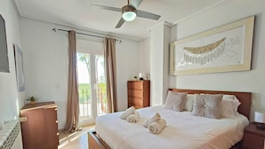 Habitación de hotel con cama, ventilador de techo, ventana con cortinas, cómoda de madera y una obra de arte enmarcada en la pared.