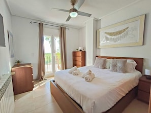 Een hotelkamer met een bed, een plafondventilator, een raam met gordijnen, een houten dressoir en een kunstwerk in een lijst aan de muur.