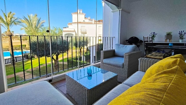 Un balcón con una mesa de vidrio, mobiliario de mimbre y vistas a una piscina y follaje.