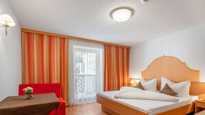 Quarto de hotel com cama de madeira, uma cadeira vermelha, uma mesinha com um vaso e uma janela com cortinas laranja.
