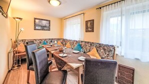 A dining area with a long table, chairs, and a patterned bench. There are two framed pictures on the walls, a floor lamp, and a window with curtains. - Lovely apartment for 6 guests with WIFI, TV, terrace and pets allowed (Mayrhofen)