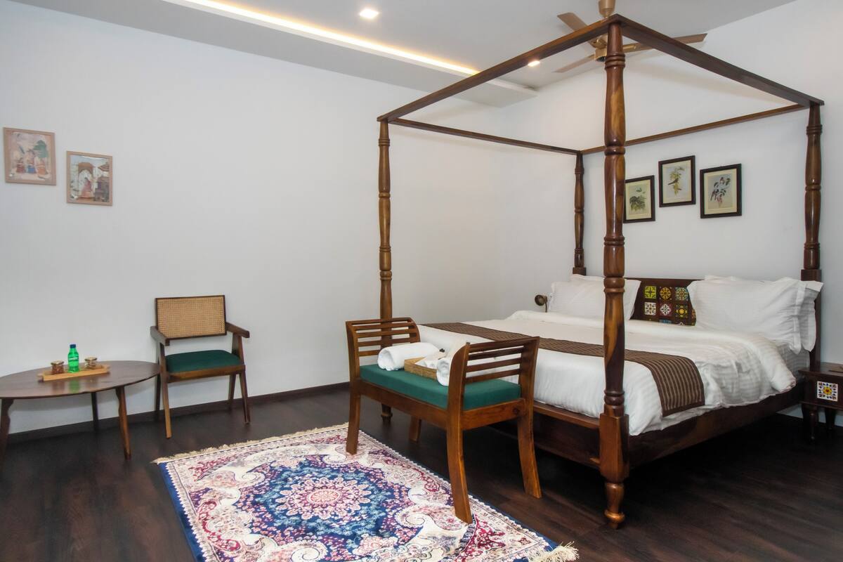 A four-poster canopy bed with a green upholstered chair and a small round table with a bottle and glasses.