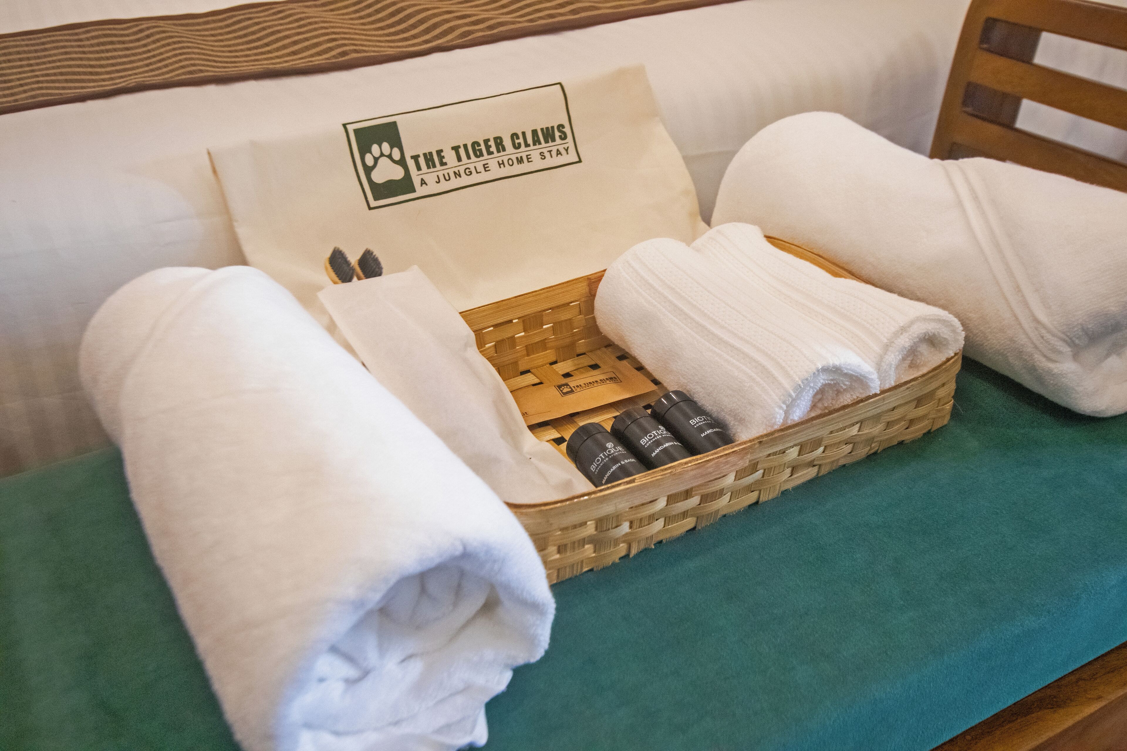 A neatly arranged bathroom with a wicker basket containing rolled towels and toiletries, a green towel on the floor, and a pillow with a logo.