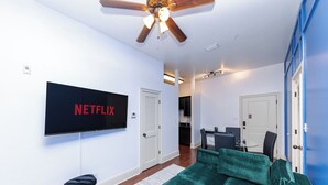 A modern living room with a green sofa, a TV displaying 'NETFLIX', a ceiling fan, and a dining area in the background.