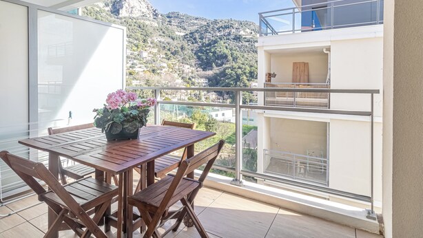 A balcony with a wooden table and chairs, a potted plant, and a view of a mountainous landscape.