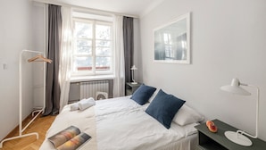 A hotel room with a bed, a bedside table, a radiator, a window with curtains, a hanging coat rack, and a wall-mounted picture. - ShortStayPoland Piwna B79 (Warsaw)