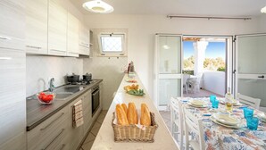 Una cocina moderna con una encimera larga, una zona de comedor con una mesa preparada para dos personas y vistas al exterior a través de una puerta de cristal.