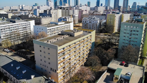 Um edifício alto com vários andares, telhado plano e área de estacionamento com vários carros.