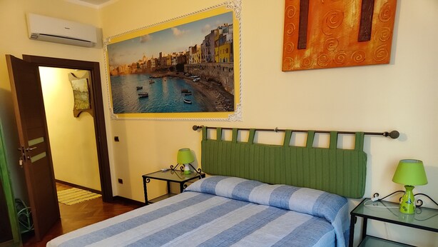 Habitación de hotel con cama, cabecera verde, cuadro de paisaje costero y una mesita con lámpara.