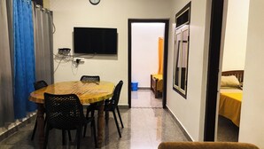Una stanza piccola e accogliente con un tavolo rotondo in legno, quattro sedie, una TV a schermo piatto montata a parete e un letto con copripiumino giallo.