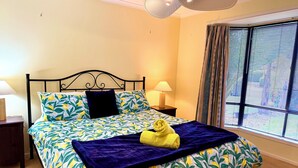 Een net opgemaakt bed met een bloemenbedekking, een donkerblauwe plaid en een gele handdoek. Er staat een nachtkastje met een lamp naast het bed.