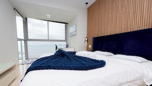 Un dormitorio con una cama grande, vista al mar, pared de paneles de madera y una lámpara moderna.
