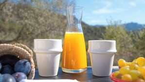 Una mesa con un vaso de jugo de naranja, dos tazas blancas, una cesta de uvas y un bol de fruta amarilla, colocada al aire libre con un cielo despejado y árboles al fondo.