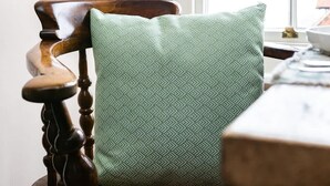 A wooden chair with a patterned cushion, a window, and a table with a lamp.