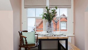 A dining area with a table set for two, a wooden chair, a vase with flowers, and a window with blinds.