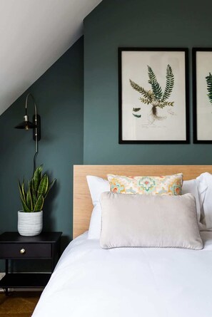 A bedroom with a bed, a nightstand with a potted plant, a wall-mounted lamp, and framed botanical prints. - Queenie | Charming 1 BR | Parking | Long stays (Ramsgate)