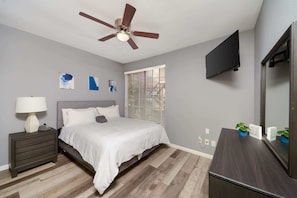 Un dormitorio con cama, mesita de noche, cómoda, ventilador de techo, televisor y ventana con persianas.