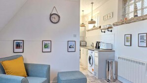 Una sala de estar moderna con un sofá, un pequeño reposapiés, un reloj, cuadros enmarcados, una zona de cocina con lavadora y un radiador.