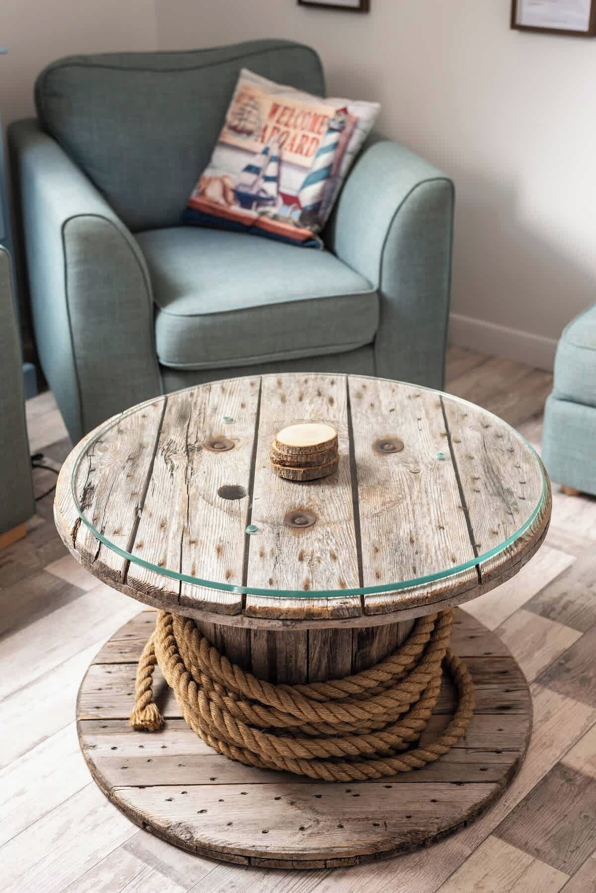 Een ronde houten tafel met een glazen blad en een touwbasis, en een kussen met een 'Welcome Aboard' ontwerp op een blauwe fauteuil op de achtergrond.