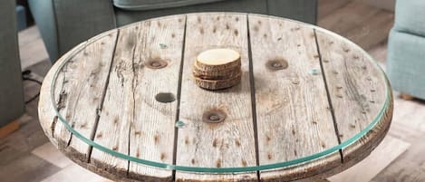 A round wooden table with a glass top, a rope base, and a cushion with a 'Welcome Aboard' design on a blue armchair in the background.