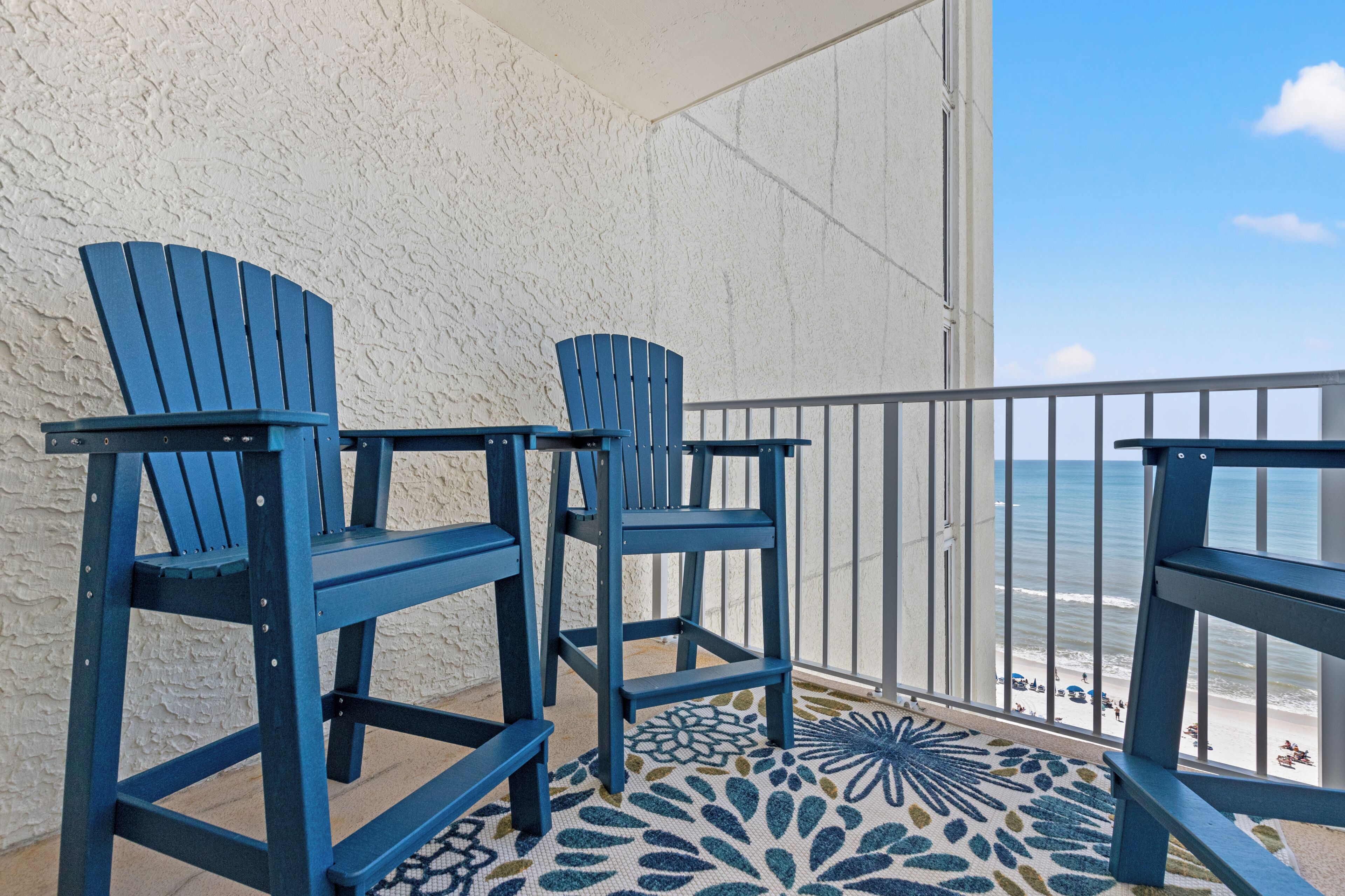 A balcony with two blue Adirondack chairs, a patterned rug, and a view of the beach.