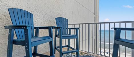A balcony with two blue Adirondack chairs, a patterned rug, and a view of the beach.