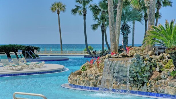 A pool with a waterfall feature, surrounded by palm trees and a view of the ocean. - Coastal Hideaway: Cozy Beachfront Studio Sleeps 4 With Stunning Views! (Panama City Beach)