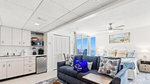 A modern living room with a grey sofa, a coffee table, and a blue bag. There is a kitchen area with a microwave and cabinets. - Coastal Hideaway: Cozy Beachfront Studio Sleeps 4 With Stunning Views! (Panama City Beach)