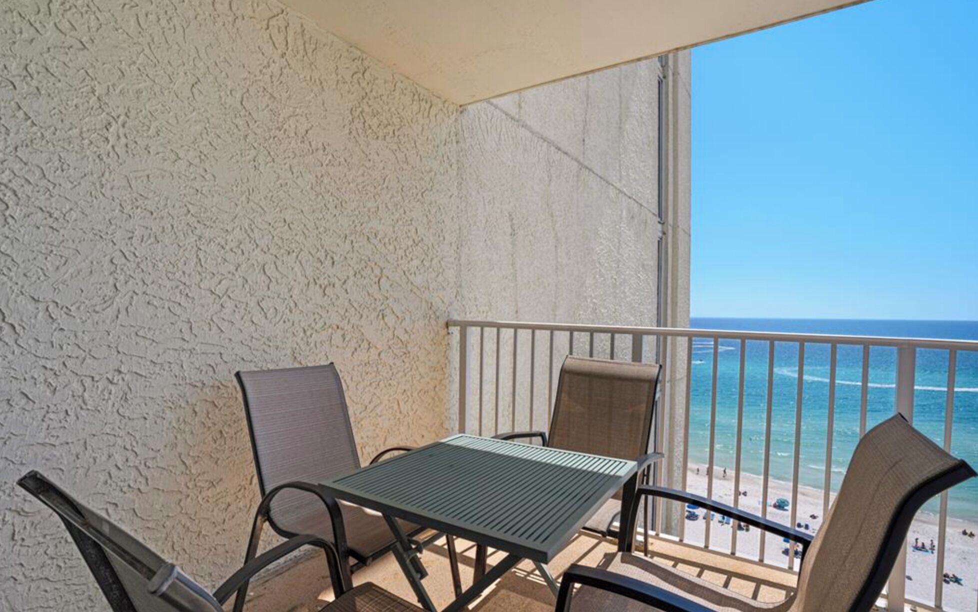 A balcony with a metal table and chairs, overlooking a beach with the ocean and clear sky.