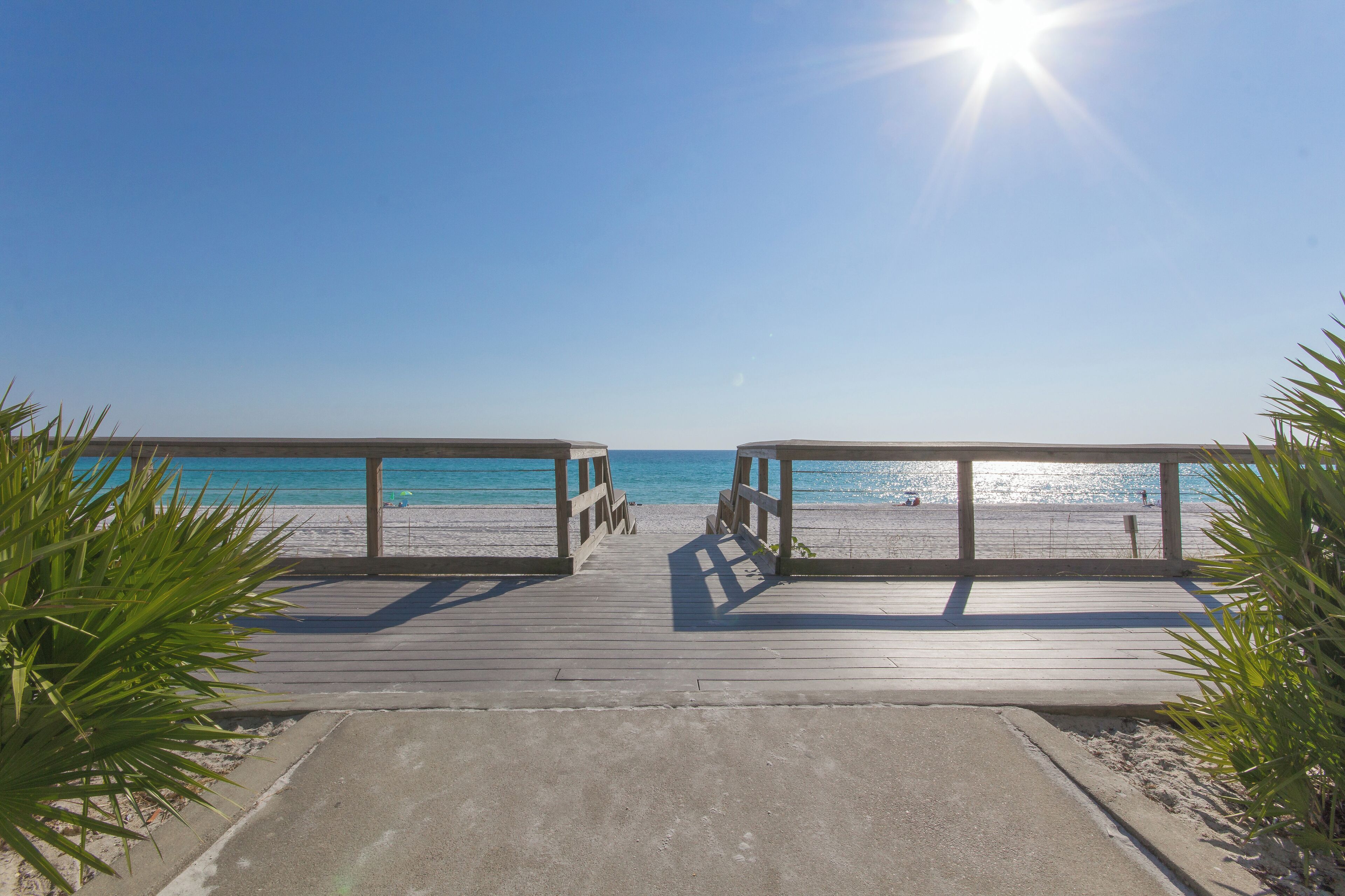 A wooden boardwalk leading to a beach with clear blue water and a bright sun in the sky.