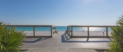 A wooden boardwalk leading to a beach with clear blue water and a bright sun in the sky.