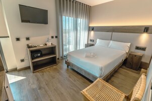 Una habitación de hotel moderna con una cama grande, un televisor de pantalla plana, un escritorio con teléfono y una mesita pequeña, y vistas a la ciudad a través de amplios ventanales.