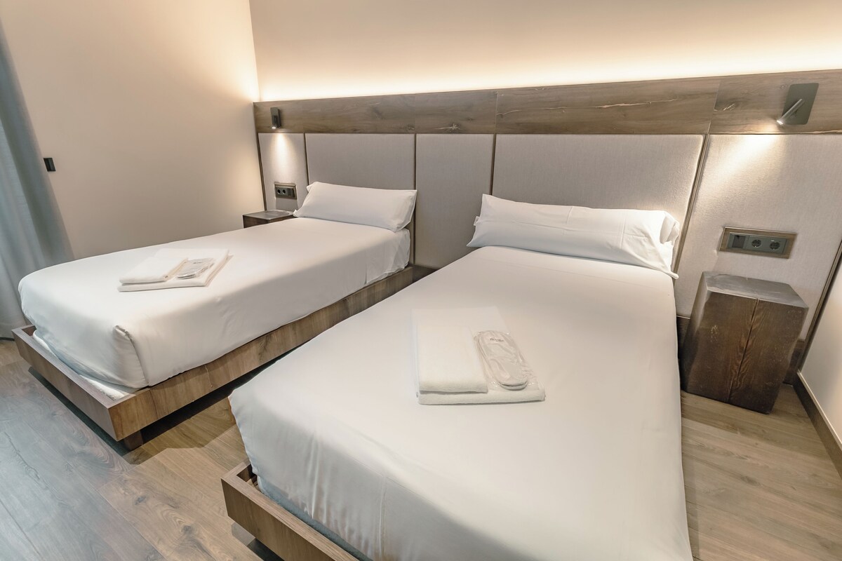 A hotel room with two beds, each with a wooden headboard and white linens. There are bedside tables with lamps and a small wooden stool.