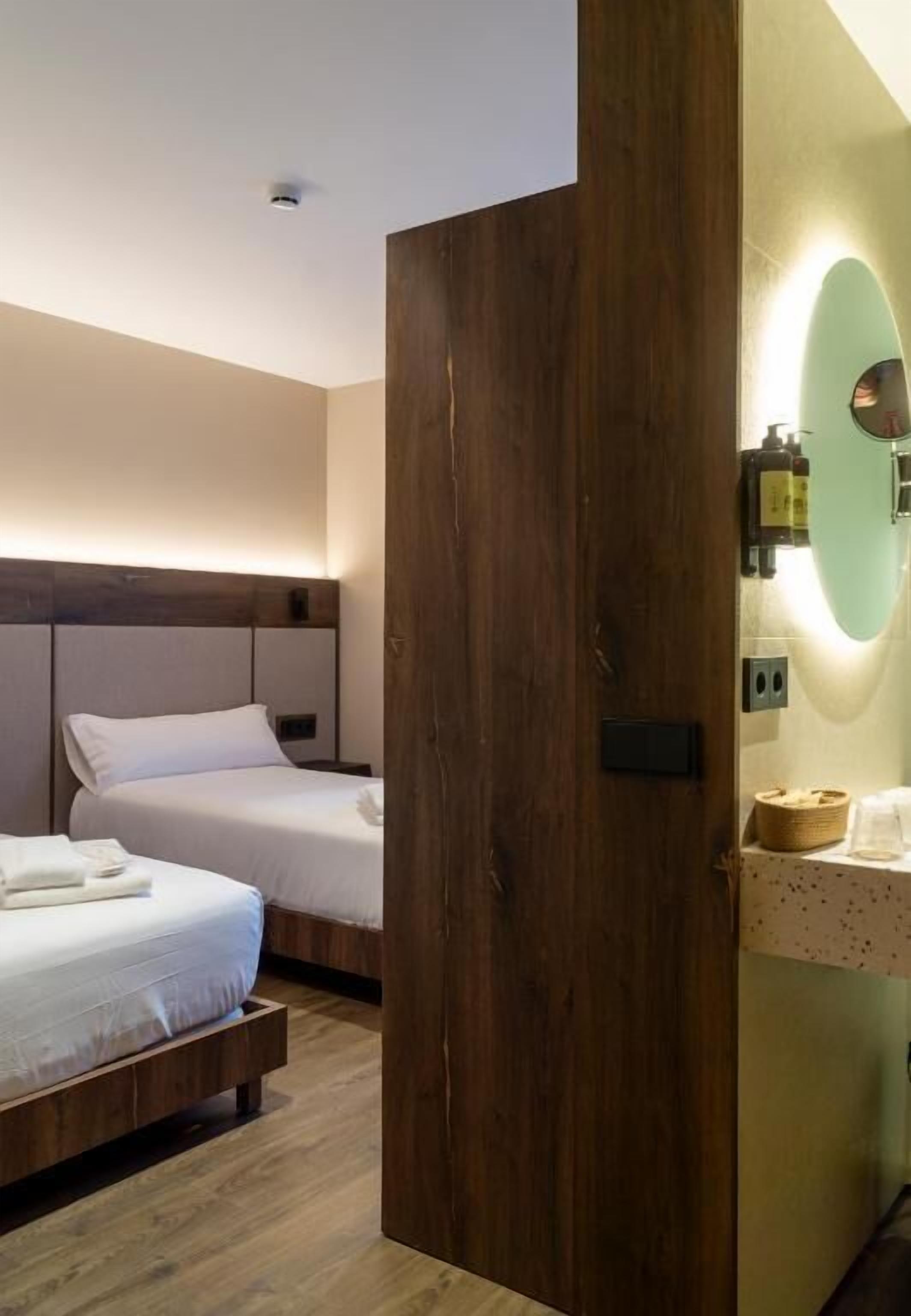 A hotel room with two beds, wooden headboards, a wooden door, a mirror, and a small shelf with toiletries.