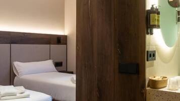 A hotel room with two beds, wooden headboards, a wooden door, a mirror, and a small shelf with toiletries.