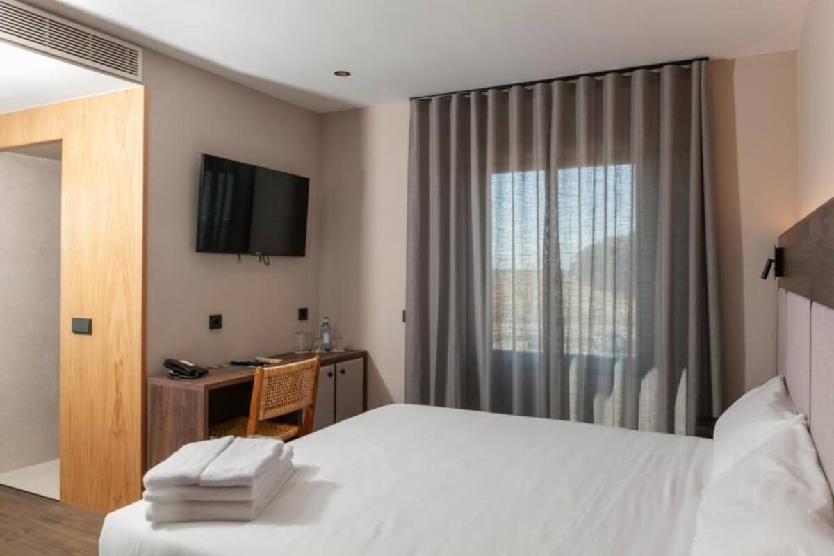 A hotel room with a bed, a desk with a telephone, a TV mounted on the wall, and a window with curtains.