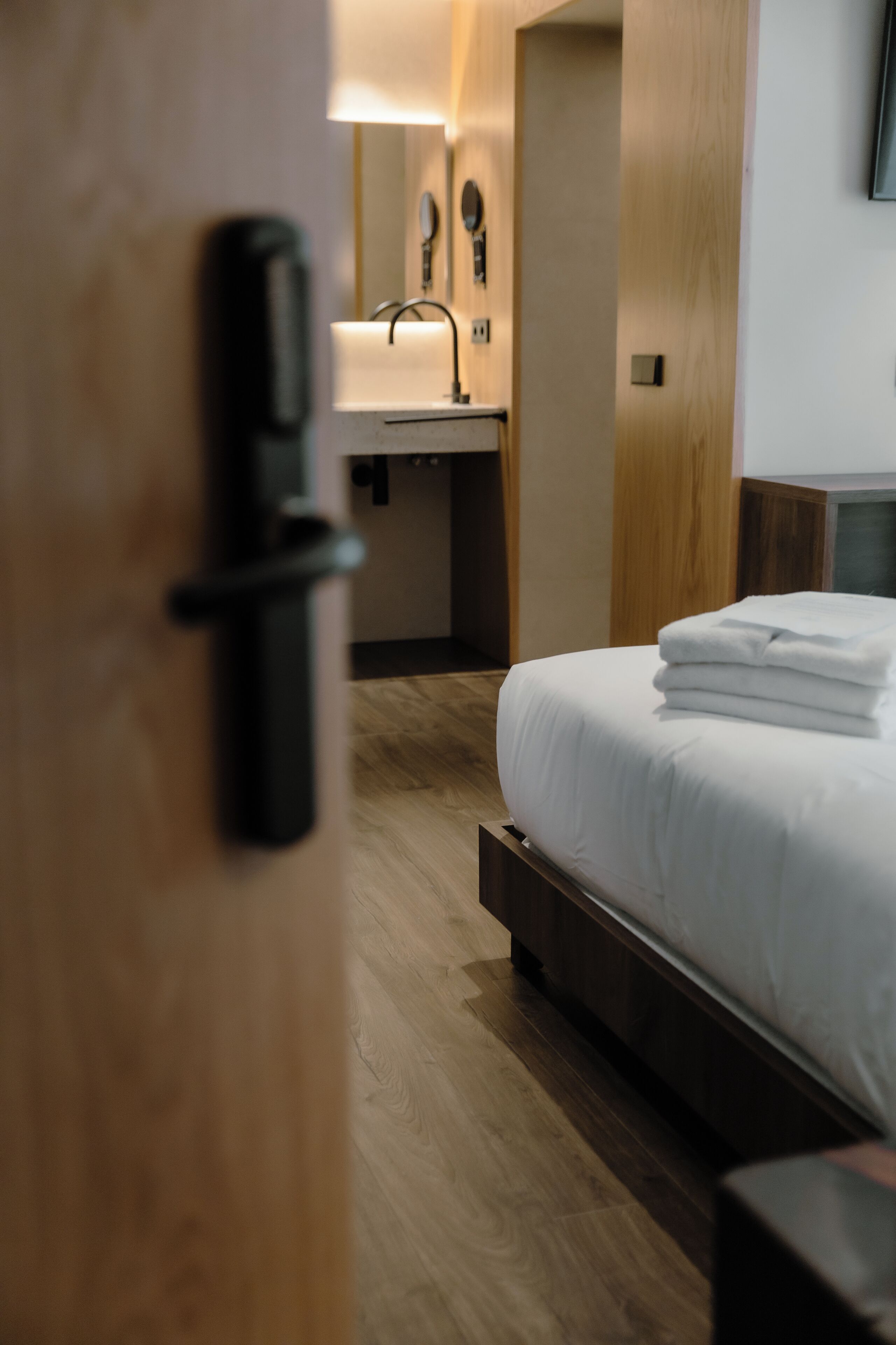 Una habitación de hotel con una cama, una mesita de noche, una lámpara y suelo de madera.