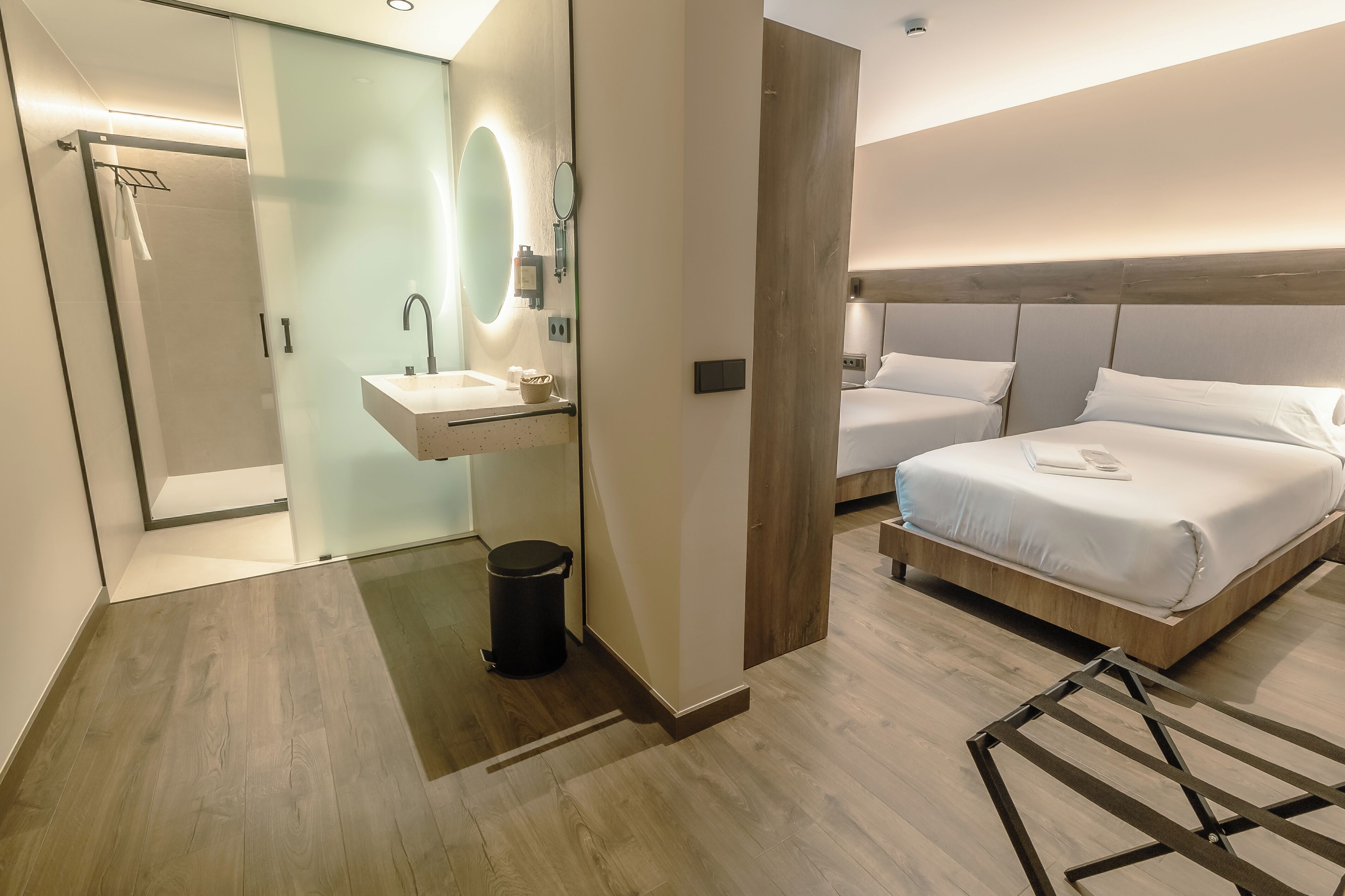 Quarto de hotel moderno com piso de madeira, box de vidro para o chuveiro, mesa de cabeceira com espelho, pia pequena e cama com roupa de cama branca.